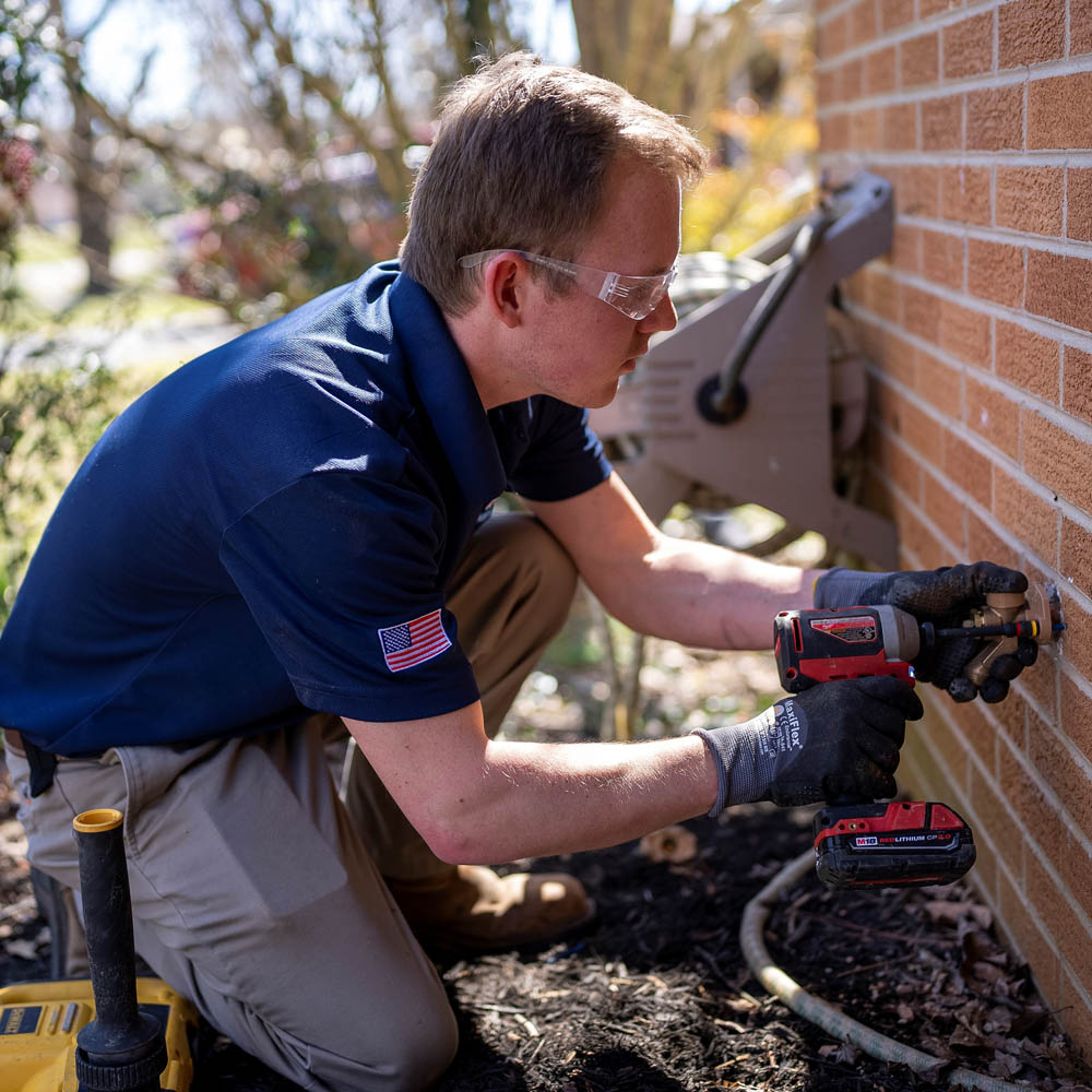 Plumber using drill in brick exterior wall of home to perform water hose spigot replacement.