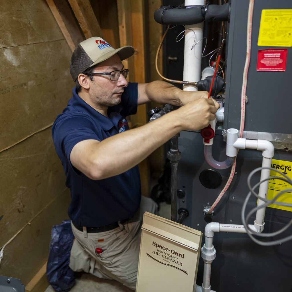 Technician in blue shirt and khakis kneeling next to the furnace he's repairing.